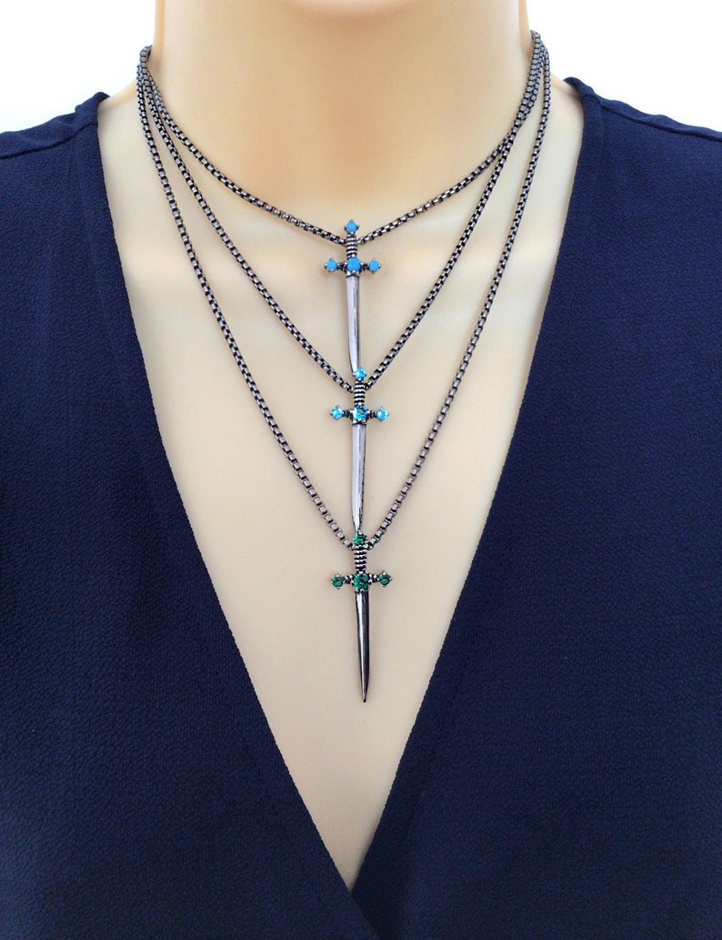 May include: Layered silver chain necklace with three sword pendants. The swords have turquoise and green gemstones.