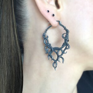 May include: A pair of silver hoop earrings with a detailed design of an octopus tentacle. The earrings are worn in a person's ear.