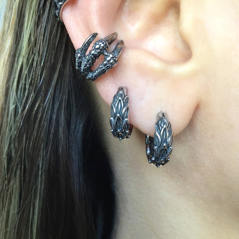 Puede incluir: Un par de pendientes de aro de plata con un dise&ntilde;o texturizado con forma de drag&oacute;n. Los pendientes se llevan en el l&oacute;bulo de la oreja.