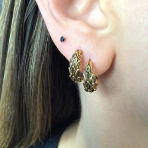 Puede incluir: Un par de pendientes de aro de oro con un dise&ntilde;o texturizado en forma de hoja. Los pendientes se llevan en los l&oacute;bulos de las orejas.