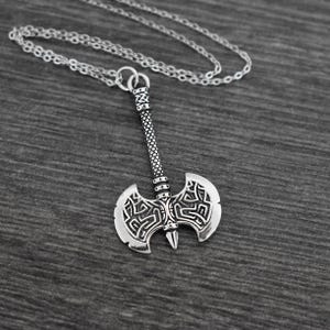 Puede incluir: Un collar de plata con un colgante de hacha detallado. El hacha tiene una hoja de doble cabeza con diseños intrincados y un mango largo.