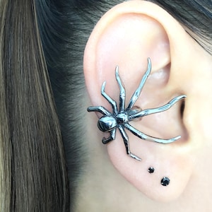 May include: A silver-colored spider ear cuff is worn on the ear. The spider has eight legs and a rounded body. Two small black studs are also visible on the earlobe. The ear is set against a background of brown hair.