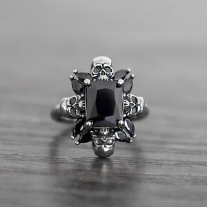 May include: A silver ring featuring a black gemstone surrounded by skull details.
