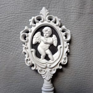 May include: White decorative key featuring a cherub design within an ornate oval frame. The cherub is in relief against a black background. The key has a detailed, scrollwork handle and frame, and is set against a gray background.