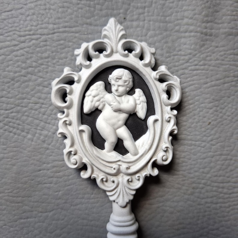 May include: White decorative key featuring a cherub design within an ornate oval frame. The cherub is in relief against a black background. The key has a detailed, scrollwork handle and frame, and is set against a gray background.