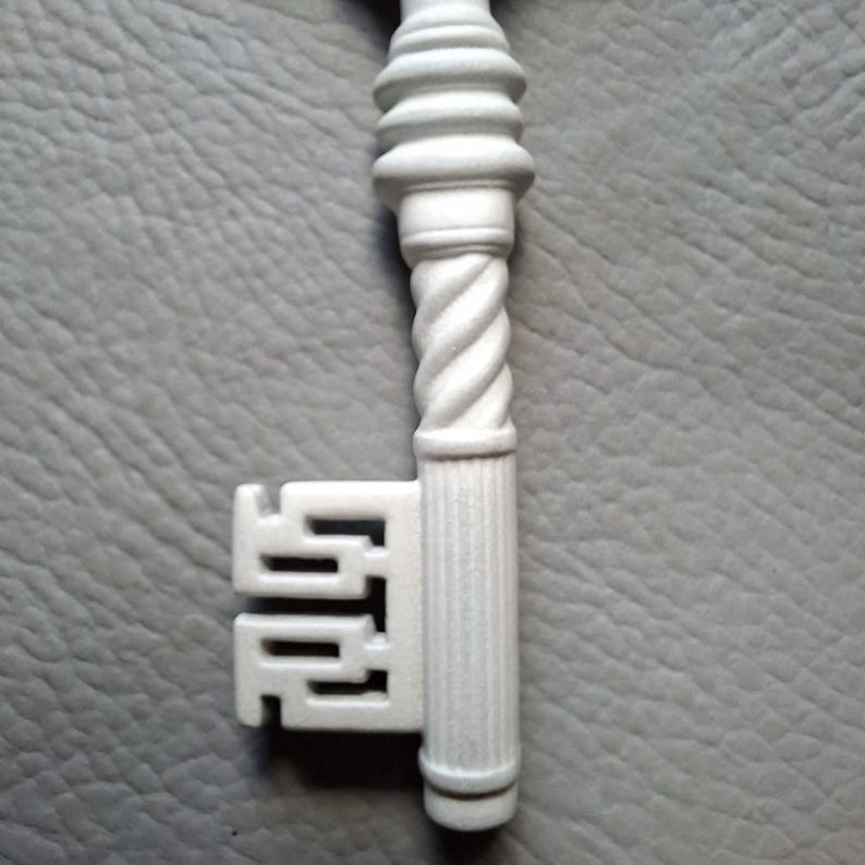 May include: A white decorative key with a detailed design. The key features a twisted stem and ornate head. The key's bit has a unique, geometric shape. The key is set against a gray background.