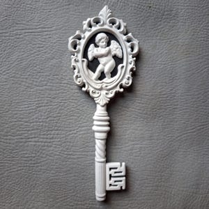 May include: White decorative key with an oval frame featuring a cherub. The key has a detailed design with a twisted handle and ornate details. The cherub is in a black oval, and the key is set against a gray background.