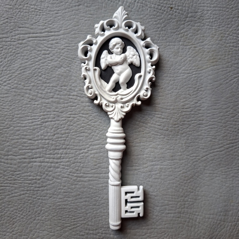 May include: White decorative key with an oval frame featuring a cherub. The key has a detailed design with a twisted handle and ornate details. The cherub is in a black oval, and the key is set against a gray background.