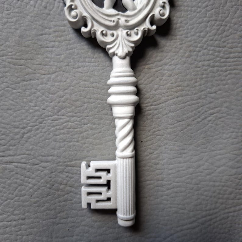 May include: A white decorative key with ornate detailing. The key features a twisted handle, a decorative top, and a detailed key bit. The key is set against a gray textured background.