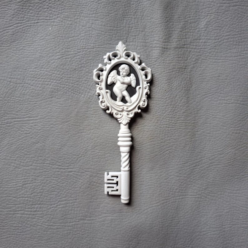 May include: A white decorative key with an oval frame featuring a cherub. The key has a detailed handle and a classic keyhole design. The background is a textured gray surface.
