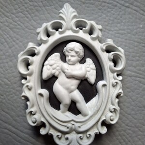 May include: A white cherub cameo in an ornate oval frame. The cherub has outstretched wings and is set against a black background. The frame is white with intricate scrollwork and decorative elements.