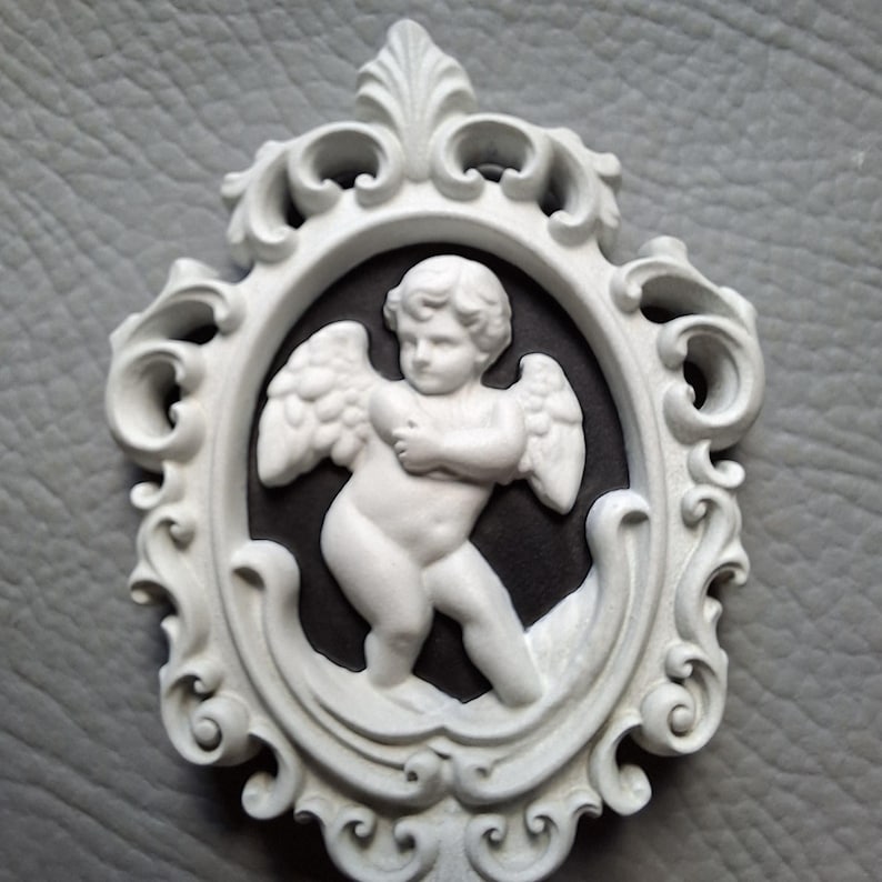 May include: A white cherub cameo in an ornate oval frame. The cherub has outstretched wings and is set against a black background. The frame is white with intricate scrollwork and decorative elements.