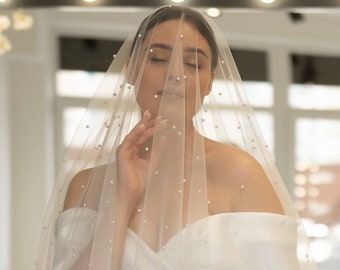 May include: A bride wearing a white off-the-shoulder wedding dress with a long veil adorned with pearl accents. The bride is standing in front of a mirror with lights.