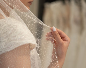 May include: A white bridal veil with a delicate pattern of small white dots and a row of pearls along the edge. The veil is made of soft, sheer fabric and is perfect for a wedding.