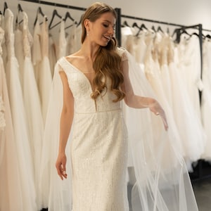 May include: A bride in a white, beaded wedding dress with a long veil, smiling as she looks at the dress selection in a bridal shop.