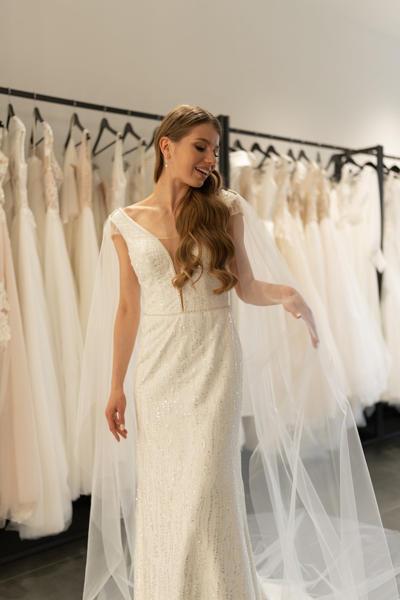 May include: A bride in a white, beaded wedding dress with a long veil, smiling as she looks at the dress selection in a bridal shop.