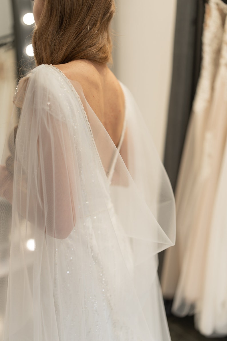 May include: A white wedding dress with a sheer, beaded cape. The dress has a low back and a fitted bodice.