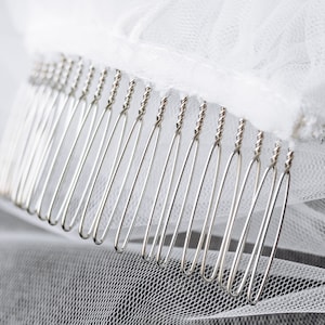 May include: A close-up of a silver comb with a row of teeth. The comb is attached to a white tulle fabric.