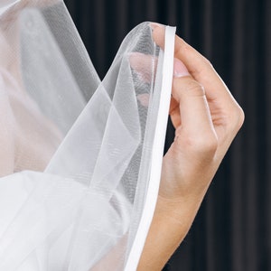 May include: A white bridal veil with a satin edge. The veil is being held by a hand.