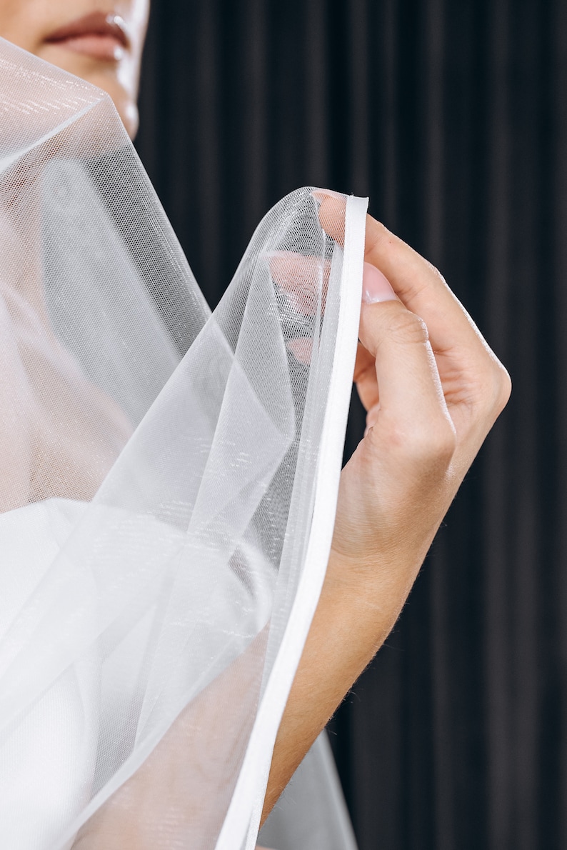 May include: A white bridal veil with a satin edge. The veil is being held by a hand.