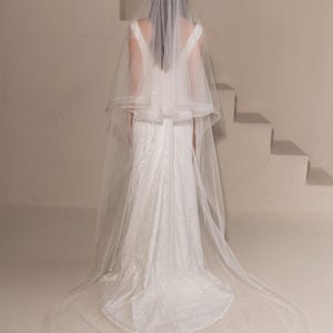 May include: A white bridal veil with a single layer of tulle and a scalloped edge. The veil is worn by a person standing in front of a white staircase.