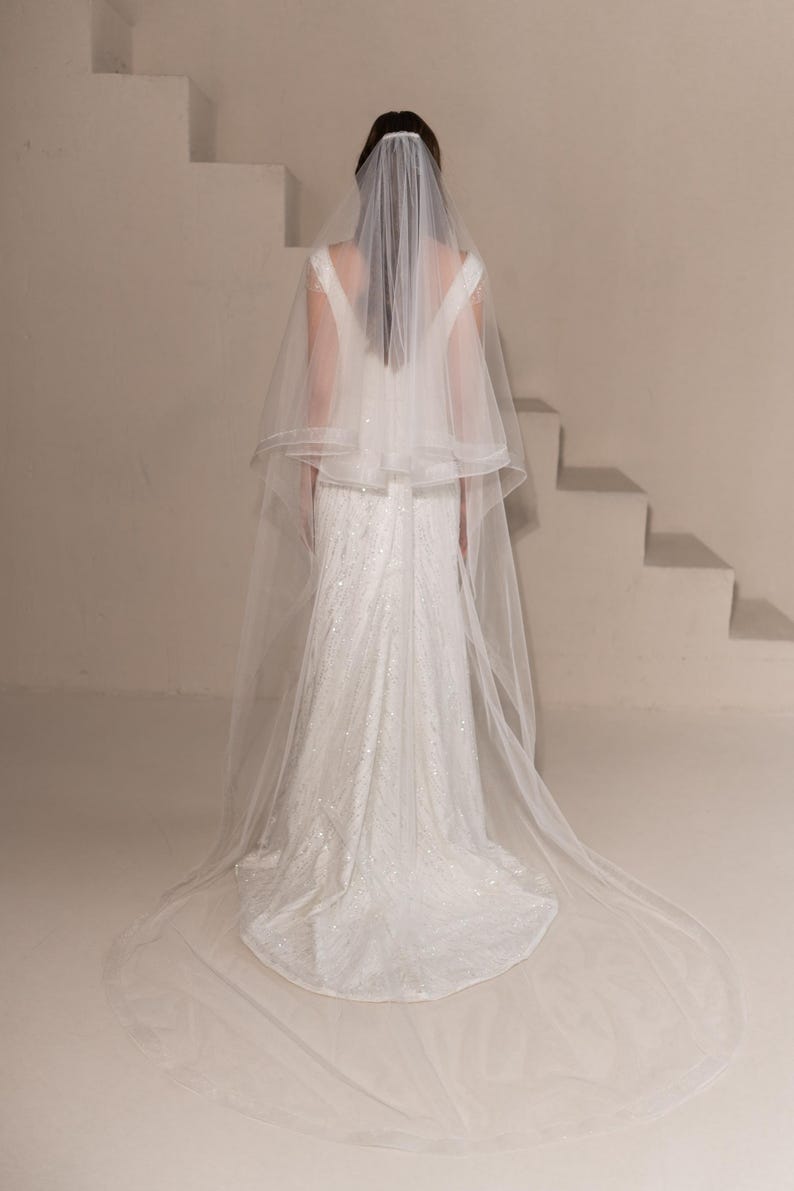 May include: A white bridal veil with a single layer of tulle and a scalloped edge. The veil is worn by a person standing in front of a white staircase.
