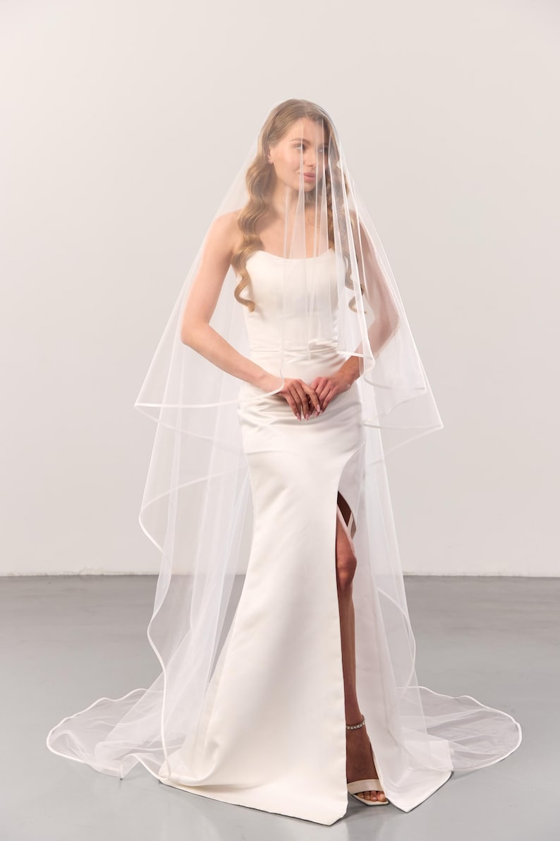 May include: A floor-length white wedding dress with a high-thigh slit and a sweetheart neckline, paired with a long, sheer, two-tier white bridal veil featuring a finished ribbon edge that drapes over the shoulders and flows to the floor.