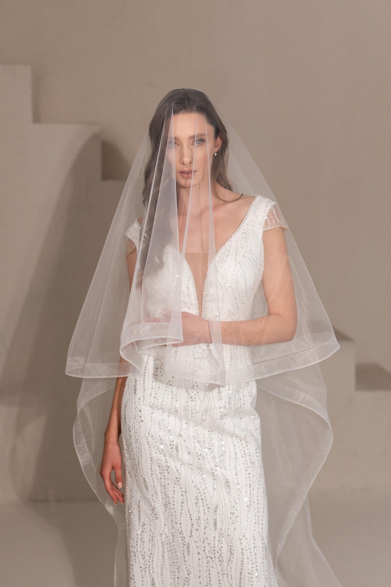 May include: A bride wearing a white wedding dress with a sequined bodice and a long veil. The veil is sheer and has a scalloped edge.
