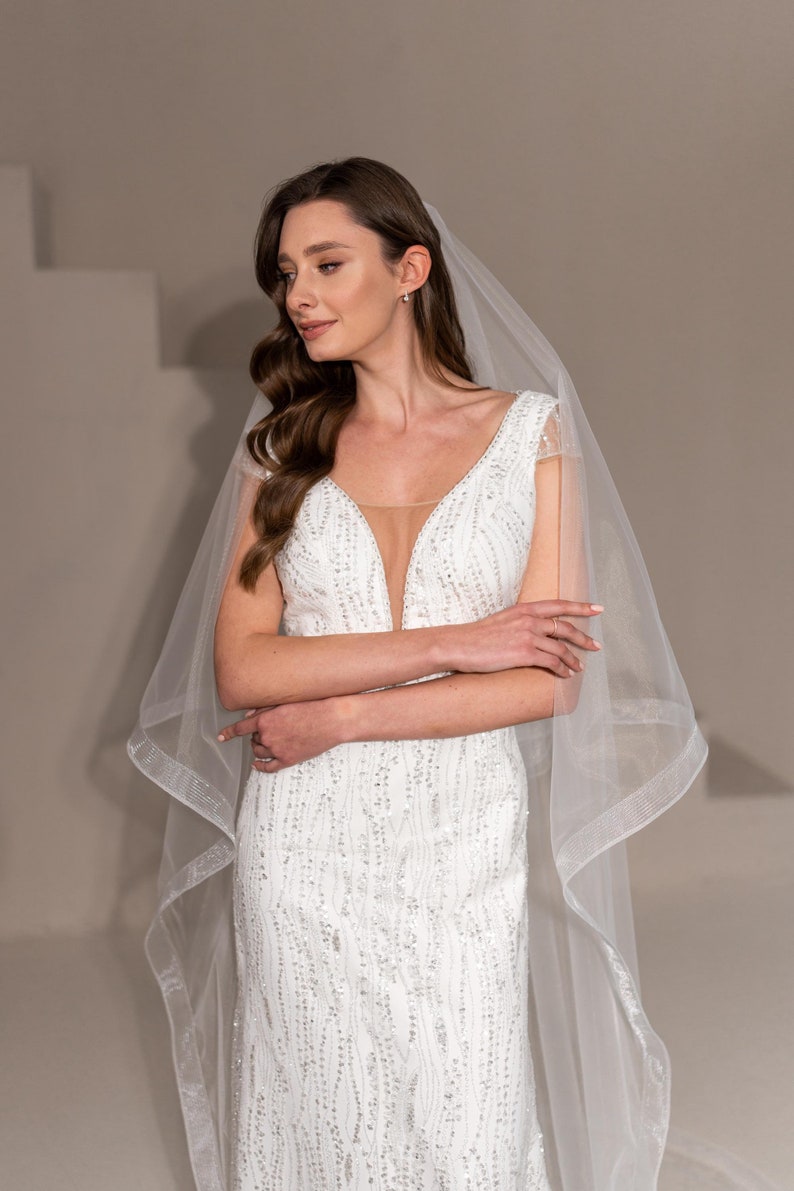 May include: A bride wearing a white, beaded, v-neck wedding dress with a long, sheer veil. The dress has a fitted bodice and a flowing skirt.