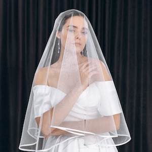 May include: A bride wearing a white wedding dress with a long, flowing veil. The veil is sheer and has a satin trim. The bride is standing in front of a black backdrop.
