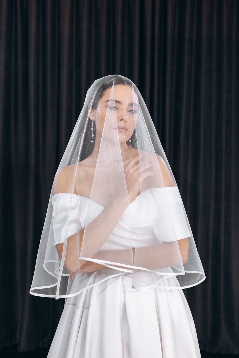 May include: A bride wearing a white wedding dress with a long, flowing veil. The veil is sheer and has a satin trim. The bride is standing in front of a black backdrop.