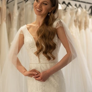 May include: A bride wearing a white, sparkly wedding dress with a sheer, flowing veil. The dress has a deep V-neckline and a fitted silhouette.