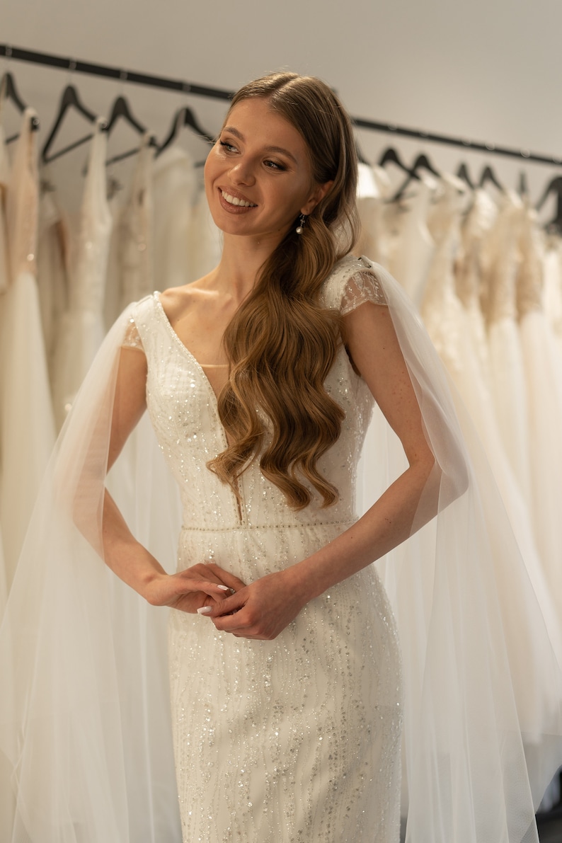 May include: A bride wearing a white, sparkly wedding dress with a sheer, flowing veil. The dress has a deep V-neckline and a fitted silhouette.