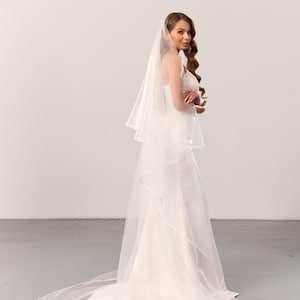 May include: A floor-length white wedding veil made of sheer tulle fabric, featuring a double-tier design with a delicate satin ribbon trim along the edges. The veil is draped over a simple white gown, extending to the floor in a circular train.
