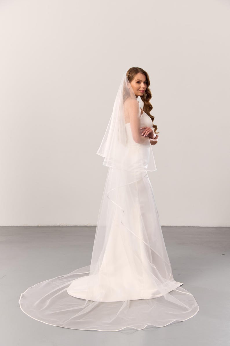 May include: A floor-length white wedding veil made of sheer tulle fabric, featuring a double-tier design with a delicate satin ribbon trim along the edges. The veil is draped over a simple white gown, extending to the floor in a circular train.