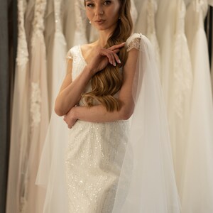 May include: A woman in a white, beaded wedding dress with a sheer veil. She is standing in front of a rack of wedding dresses.