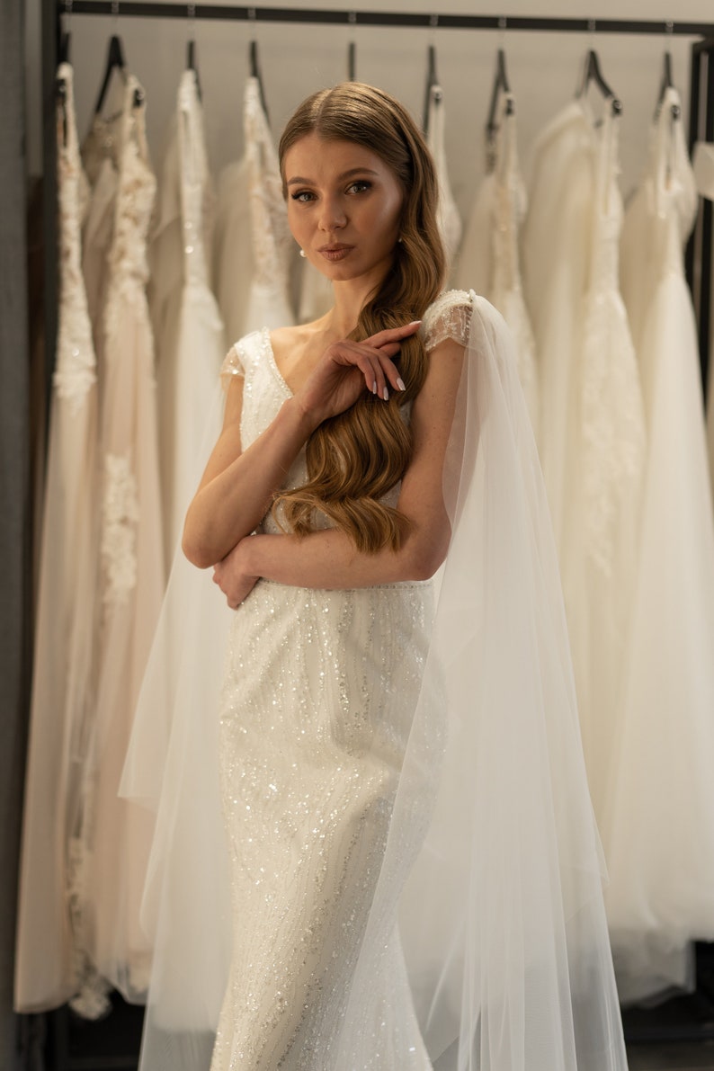 May include: A woman in a white, beaded wedding dress with a sheer veil. She is standing in front of a rack of wedding dresses.