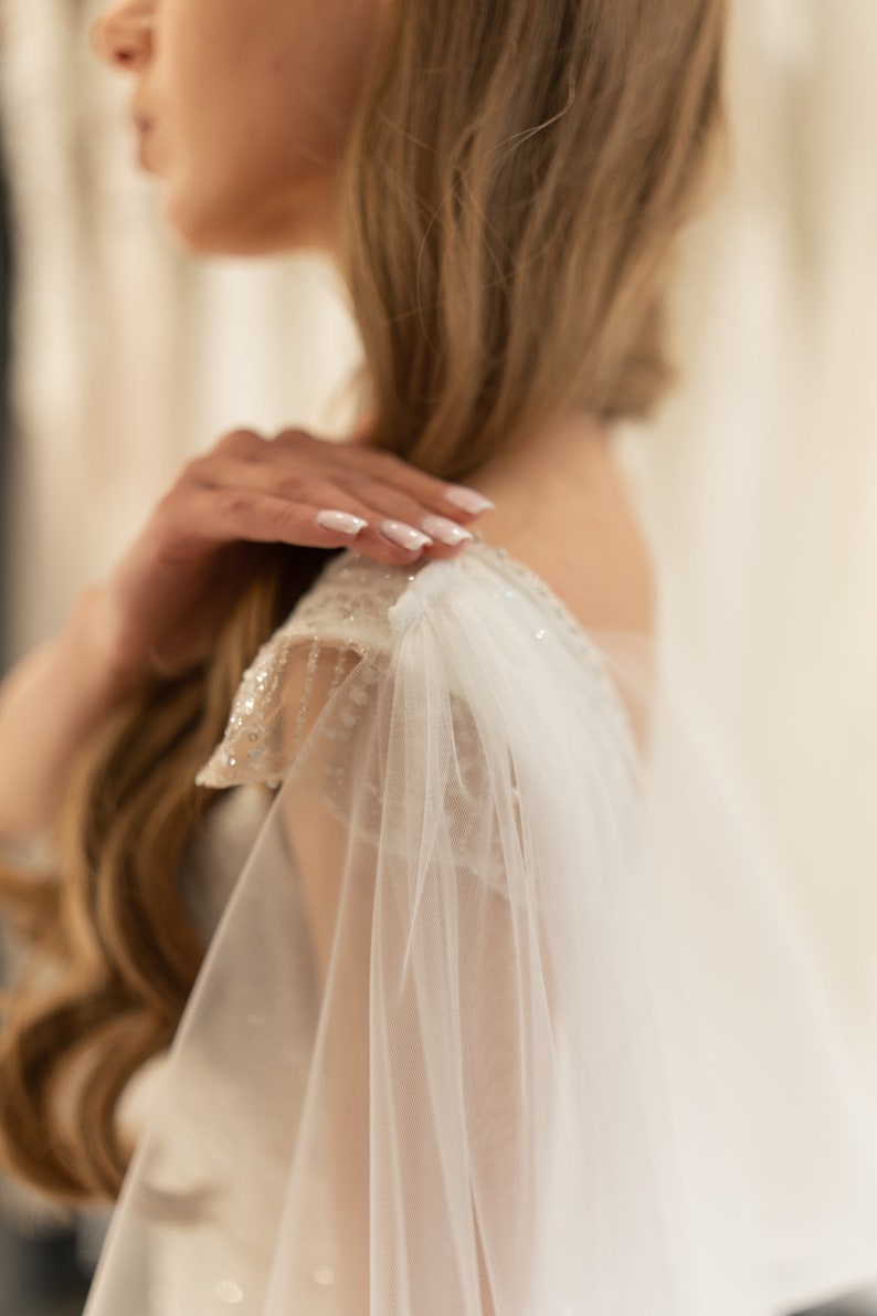 May include: A white bridal veil with delicate lace detailing. The veil is draped over the shoulder of a person wearing a white dress.