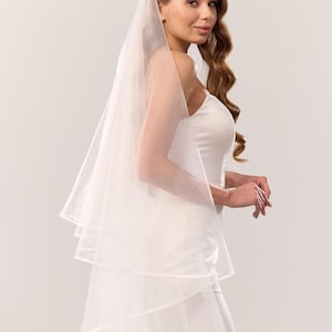 May include: A white multi-layered tulle wedding veil featuring a clean, finished edge. The sheer, lightweight fabric drapes elegantly over the shoulders, creating a soft, layered effect that complements the strapless white gown worn underneath.