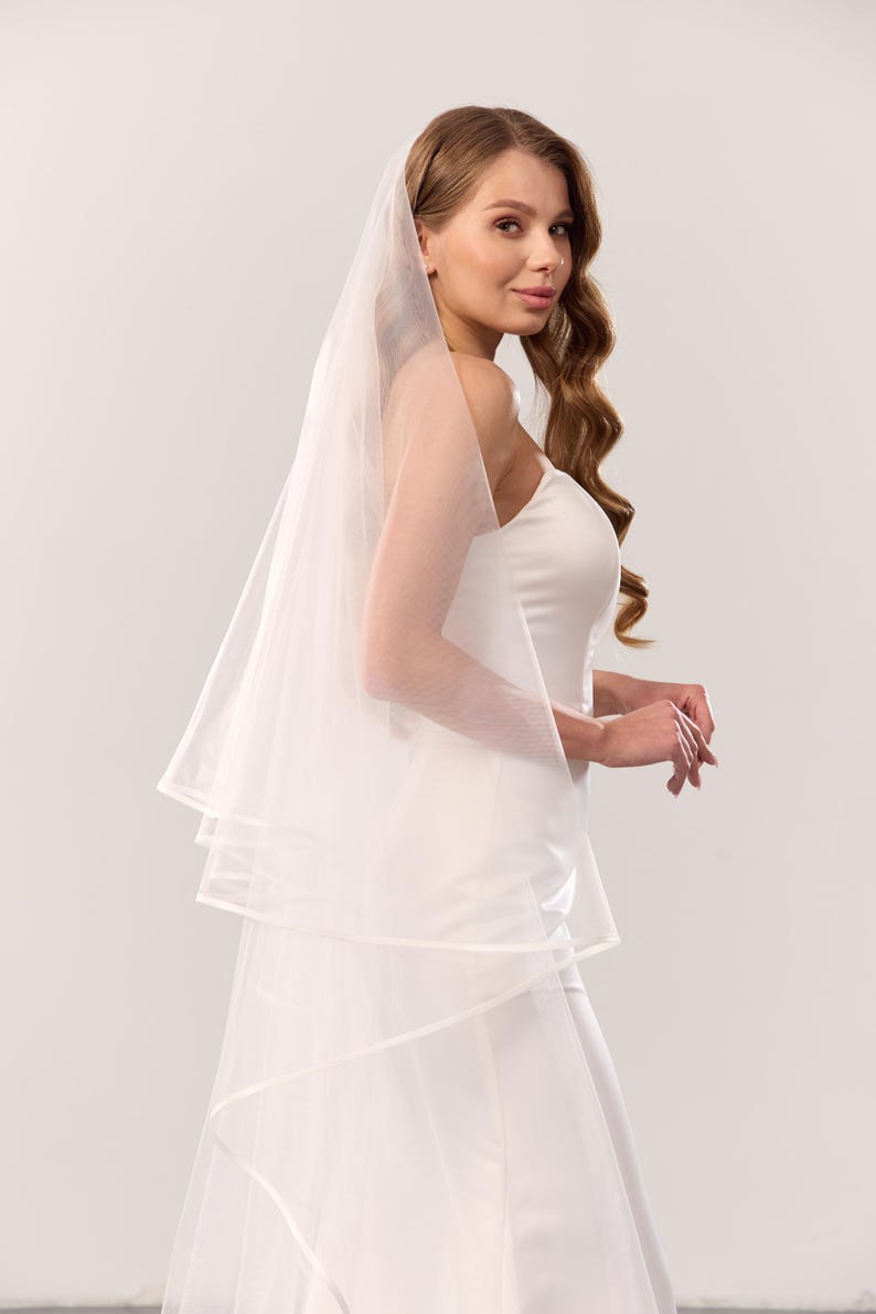 May include: A white multi-layered tulle wedding veil featuring a clean, finished edge. The sheer, lightweight fabric drapes elegantly over the shoulders, creating a soft, layered effect that complements the strapless white gown worn underneath.