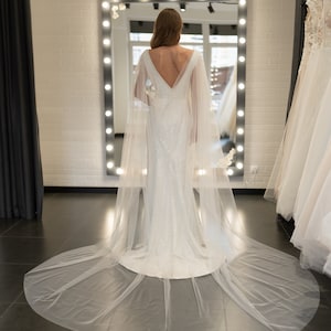 May include: A white wedding dress with a long train and a sheer cape. The dress has a fitted bodice and a flowing skirt. The bride is standing in front of a mirror.