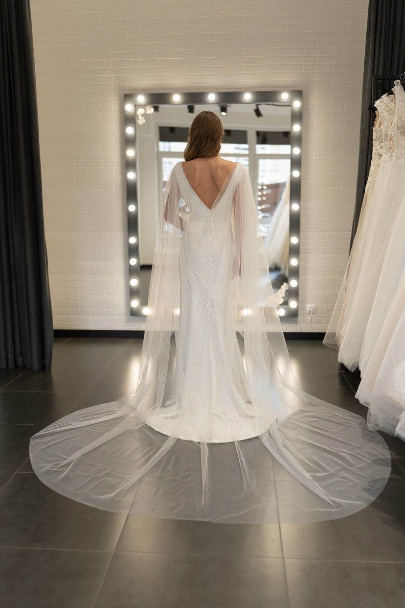 May include: A white wedding dress with a long train and a sheer cape. The dress has a fitted bodice and a flowing skirt. The bride is standing in front of a mirror.