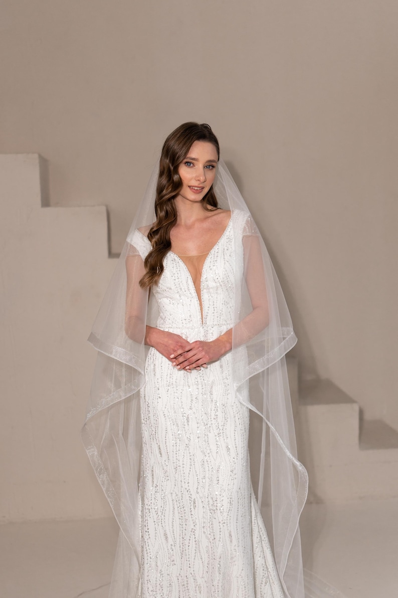 May include: A bride wearing a white, beaded, v-neck wedding dress with a long, flowing veil. The dress has a fitted bodice and a flowing skirt.