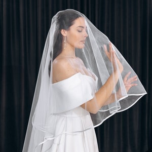 May include: A bride wearing a white wedding dress with a long, flowing veil. The veil is made of tulle and has a satin trim. The bride is standing in front of a black backdrop.