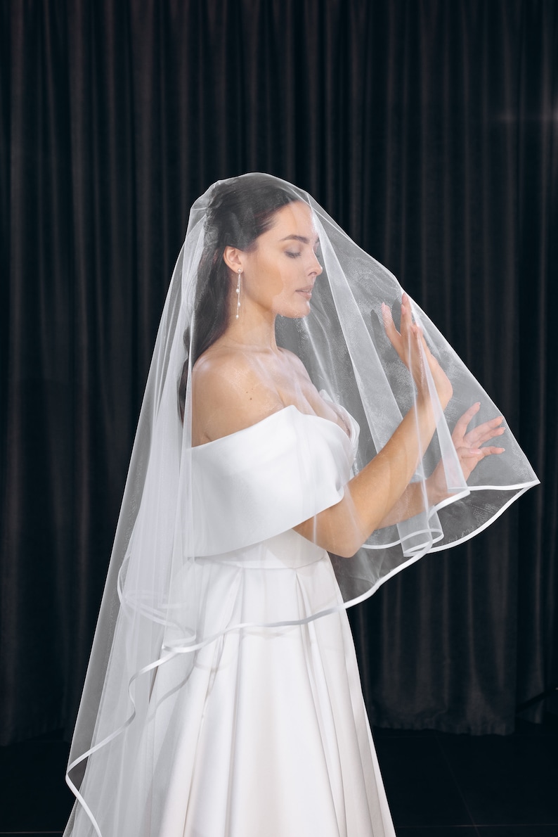 May include: A bride wearing a white wedding dress with a long, flowing veil. The veil is made of tulle and has a satin trim. The bride is standing in front of a black backdrop.
