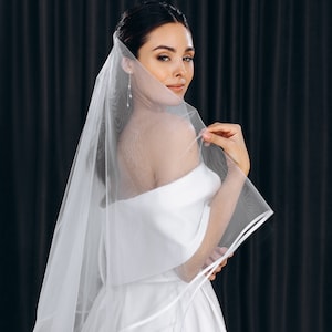 May include: A bride wearing a white off-the-shoulder wedding dress with a long, flowing veil. The veil has a satin trim and is draped over the bride's shoulders.