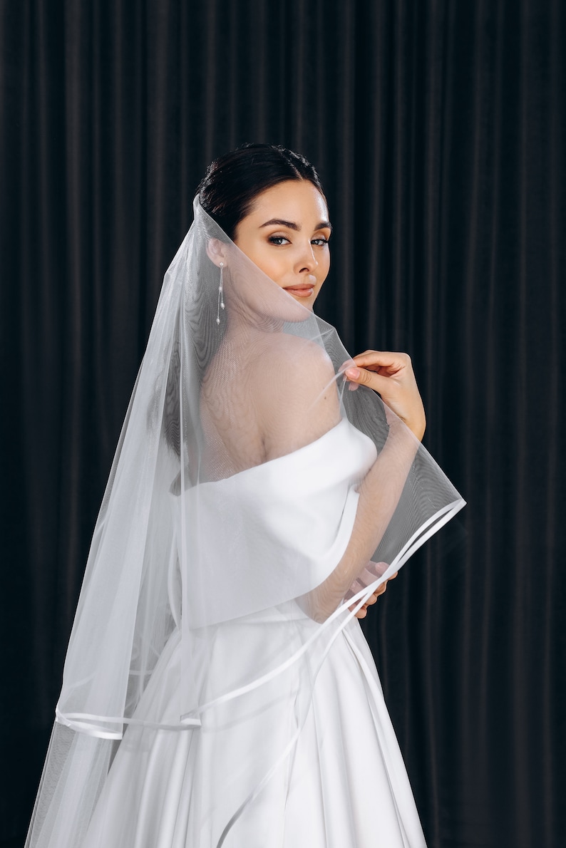 May include: A bride wearing a white off-the-shoulder wedding dress with a long, flowing veil. The veil has a satin trim and is draped over the bride's shoulders.