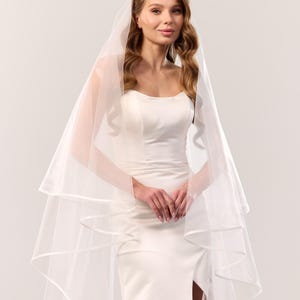 May include: A white wedding veil featuring two tiers of sheer tulle fabric with a satin ribbon trim along the edges. The veil is draped over the head and shoulders, cascading down in soft layers, paired with a sleeveless white floor-length dress.