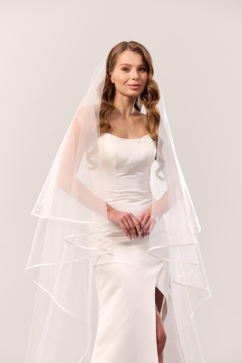 May include: A white wedding veil featuring two tiers of sheer tulle fabric with a satin ribbon trim along the edges. The veil is draped over the head and shoulders, cascading down in soft layers, paired with a sleeveless white floor-length dress.