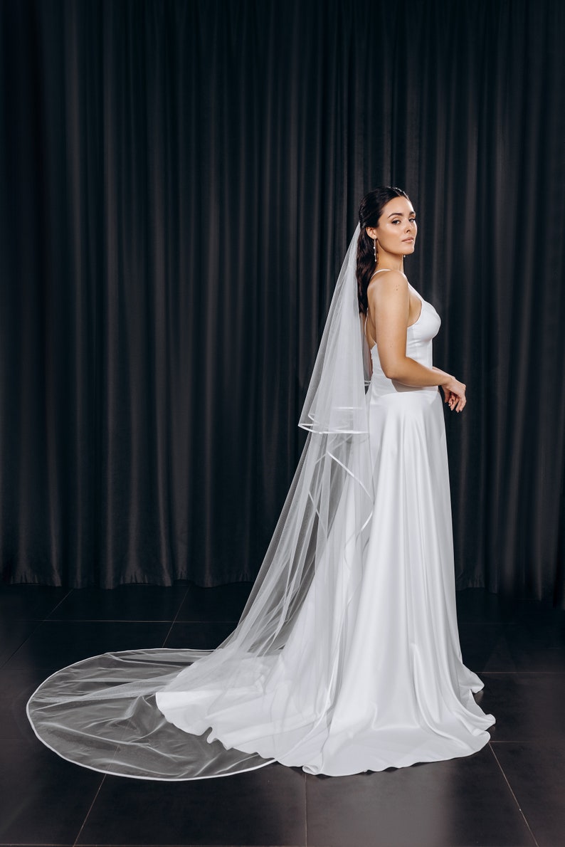 May include: A bride wearing a white wedding dress and a long, white veil with a satin trim. The veil is flowing behind her and she is standing in front of a black backdrop.